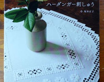 Op de afbeelding: Een wit geborduurd kleed met een bloemmotief en een kleine vaas met een roos. De boekomslag op de achtergrond heeft Japanse tekst. Het kleed ligt op een houten oppervlak.