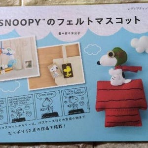 Puede incluir: Una portada de libro con una casa de perro de fieltro rojo con un perro de dibujos animados que lleva un gorro de piloto y gafas. El título del libro es "SNOOPY™'s Felt Mascot" y también se puede ver el texto "52 items to make".