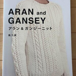 Op de afbeelding: Een boekomslag met een crèmekleurige Aran-trui met ingewikkelde kabelbreipatronen. De titel "ARAN and GANSEY" staat in grote, vette zwarte letters, met Japanse tekst eronder. De trui wordt door een persoon gedragen.