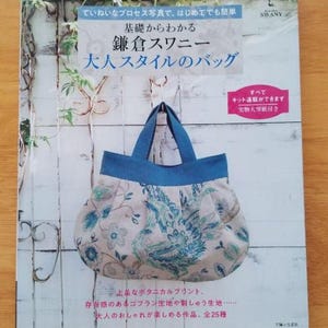 May include: A book cover featuring a floral patterned handbag with blue handles. The book title is in Japanese, with text indicating it's about making bags. The background is a light wood surface.