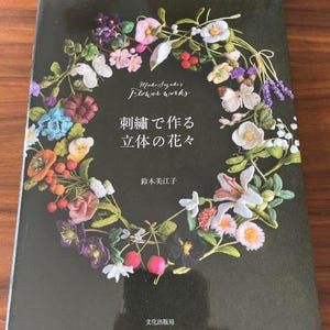 Könnte beinhalten: Ein Buchcover mit dunkelgrünem Hintergrund zeigt einen floralen Kranz. Der Kranz besteht aus verschiedenen bunten gestickten Blumen, Schmetterlingen und Blättern. Der Titel "Michiko Suzuki's Flower Works" steht über japanischem Text.