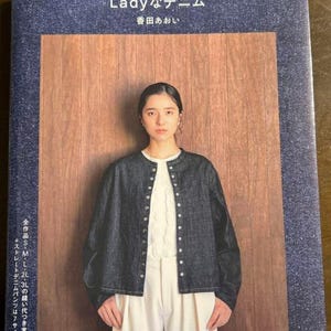 Puede incluir: Portada de libro con texto japonés "Lady Denim" y nombre del autor. La portada presenta a una mujer con una chaqueta vaquera oscura sobre una camiseta y pantalones blancos. El fondo es un panel de madera.