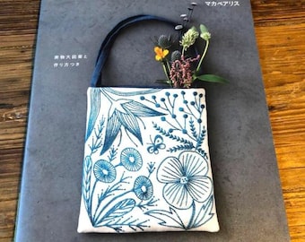 May include: A book titled "Botanical Embroidery: Plant Embroidery Handbook" with a blue cover and a white tote bag with blue embroidery of flowers and leaves. The tote bag has a blue handle and is sitting on top of the book.