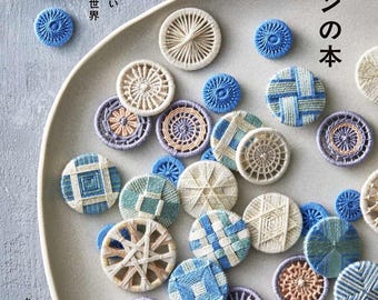 Puede incluir: Colección de botones hechos a mano en varios diseños y colores, incluyendo azul, blanco y beige. Los botones están dispuestos sobre un plato gris, con texto japonés visible. Los botones están hechos con hilo y presentan patrones intrincados.