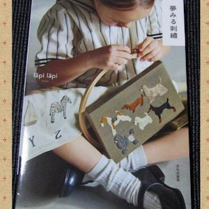 以下が含まれることがあります： 「愛らしい刺繍」というタイトルの本。女の子が刺繍された動物がついた茶色とベージュのバッグを持っている。バッグは木製のハンドルで、女の子はストライプのシャツと白いズボンを履いている。