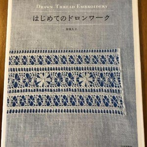 Peut inclure: Un livre intitulé "Drawn Thread Embroidery:  Beginning Drawn Thread Work" par  著者:  久米久子. La couverture présente un motif brodé blanc sur un fond en tissu bleu.