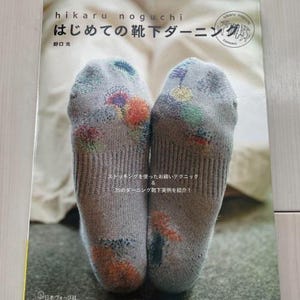 Puede incluir: Un libro titulado "はじめての靴下ダーニング" (Hajimete no Kutsuta Darning) de Hikaru Noguchi, que muestra un primer plano de dos calcetines bordados de colores.