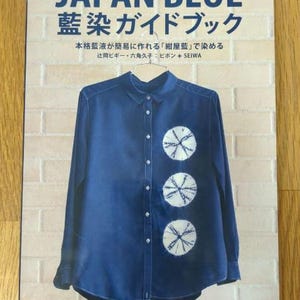 Op de afbeelding: Een boek getiteld "JAPAN BLUE 藍染ガイドブック" met een blauw shirt met witte cirkels. De tekst op de omslag luidt "本格藍液が簡易に作れる「紺屋藍」で染める 辻岡ビギー・六角久子: ピポン + SEIWA"