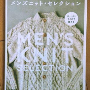 May include: A book cover featuring a cream-colored knit cardigan with wooden buttons. The title "MEN'S KNIT SELECTION" is prominently displayed in large white letters. Japanese text is at the top.