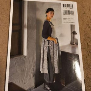 Pu&ograve; includere: Un grembiule a quadri bianchi e neri con una maglia nera e pantaloni neri. Il grembiule ha un laccio in vita e una tasca sul davanti. La modella &egrave; in piedi in una stanza con un pavimento in legno e una finestra sullo sfondo.