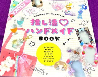May include: A colorful book titled "OSHIKATSU HANDMADE BOOK" with Japanese text and illustrations of handmade crafts. The cover features a pink tote bag, a heart-shaped keychain, and various decorative items. The background is a purple surface.