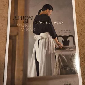 Pu&ograve; includere: Un libro intitolato "Apron and Work Wear" che mostra una donna che indossa un grembiule bianco e una camicetta nera. La copertina del libro include anche il testo "エプロンとワークウェア" in giapponese.