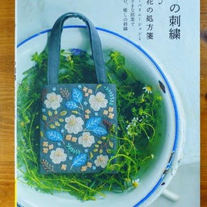 May include: A book titled "yula no  ori  zushi" with a blue and white tote bag with floral embroidery on the cover. The book is on a wooden surface.