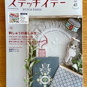 May include: A Japanese embroidery magazine titled "Stitch Idées" with a cover featuring a blue and white embroidered design, a gray cat, and a pincushion with a black and white checkered pattern.