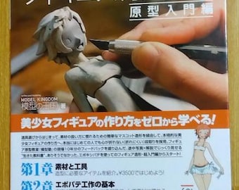 May include: A Japanese language book titled "How to build GARAGE KIT Vol. 1: Figure Textbook, Original Entry Edition" with a cover image of a person sculpting a female figure with a sculpting tool. The book cover also features a cartoon illustration of a woman in a bikini.