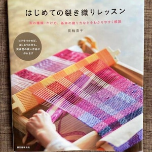 Op de afbeelding: Een boek getiteld "はじめての裂き織りレッスン" (Beginnersles voor weven) met een kleurrijke geweven stof op de omslag. De omslag van het boek toont een close-up van een hand die weeft op een weefgetouw.