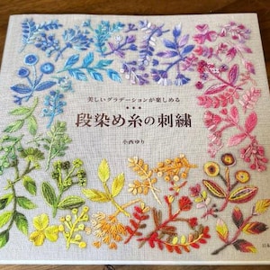 Könnte beinhalten: Ein Buch mit dem Titel "段染め糸の刺繍" (Stickerei mit Verlaufsgarnen) von Yuri Konishi. Der Umschlag zeigt ein farbenfrohes Stickmuster mit verschiedenen Blumen und Blättern.