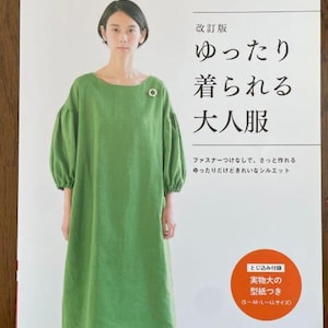 May include: A woman wearing a green dress with a round neckline and puffed sleeves. The dress has a small brooch. The cover of a Japanese book is visible with text in Japanese.