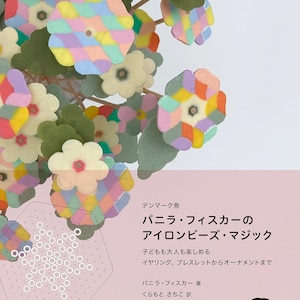 Puede incluir: Una portada de libro con fondo rosa y una ilustración colorida de una flor hecha de cuentas de hierro. El título del libro es "La magia de las cuentas de hierro de Pernille Fisker".