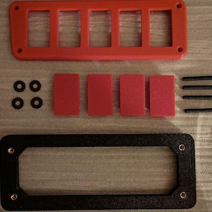 May include: An assortment of components for an electronic device, including a red rectangular panel with five openings, four red rectangular inserts, a black rectangular frame, black screws, and black washers. The parts are arranged on a light-colored surface.