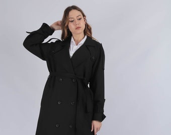 May include: A black double-breasted trench coat featuring a wide lapel, shoulder epaulets, long sleeves with buckled cuffs, and a matching waist belt. The coat is worn over a white collared shirt, blue denim jeans, and white sneakers.