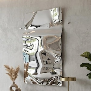 Può includere: Un grande specchio argentato astratto è appeso a una parete grigio chiaro. Sotto, un mobile in legno scuro con dettagli dorati espone libri e oggetti decorativi. Una poltrona color crema con una coperta e una pianta in vaso completano l'interno moderno.