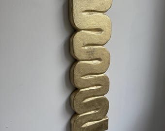 May include: A gold-coloured, wavy sculpture mounted on a white wall. The sculpture has a textured surface and a unique, abstract design. The piece is a decorative item, likely for interior design.