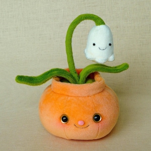 May include: A plush orange flower pot with a smiling face, holding a green stem with a white flower. The flower pot has large, round eyes and a small pink nose. The white flower has a smiling face.