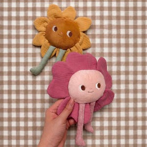 May include: Two plush flower toys with embroidered faces. One is a golden-yellow sunflower with a brown center and light green stem. The other is a magenta pink flower with a light pink center and matching pink stem, held by a hand.