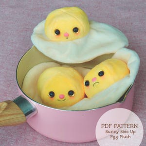 Puede incluir: Una sartén rosa contiene tres peluches de huevos fritos. Los huevos tienen yemas amarillas, claras de huevo blancas y caras bonitas de dibujos animados con ojos negros y mejillas rosas. El texto "PDF PATTERN Sunny Side Up Egg Plush" está en la esquina inferior derecha.