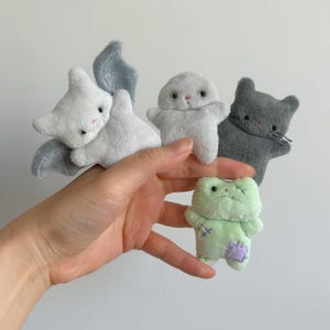May include: A hand displays five small plush toys. The toys include a white bat with grey wings, a grey bunny, a grey cat, and a light green frog with a purple patch. The toys have black eyes and pink accents.