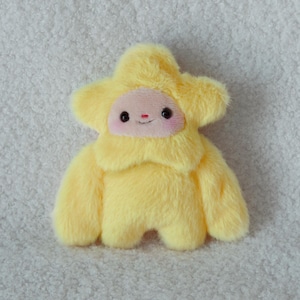 May include: A plush toy shaped like a star with bright yellow faux fur. The center features a smooth, light pink face with two small black eyes, a tiny red nose, and a thin stitched smile. The star has five points and two short arms.