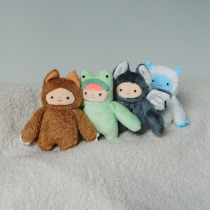 May include: Four plush dolls in animal costumes: a brown bear, a green frog, a gray bunny, and a blue yeti. Each doll has a simple face with a small mouth and two dots for eyes. The dolls are arranged in a row on a white, textured surface.