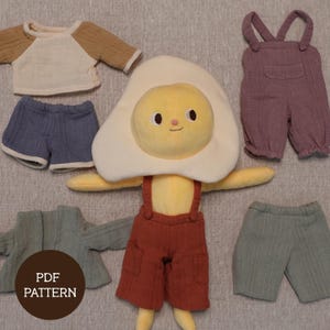 May include: A plush toy shaped like a fried egg with various outfits. The outfits include a brown and cream raglan shirt, blue shorts, purple overalls, a green jacket, and green shorts. The text "PDF PATTERN" is on a brown circle.