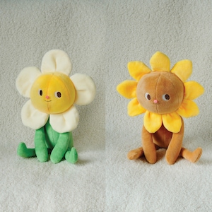 May include: Two plush flower toys. One has a yellow centre, white petals, and green stem and leaves. The other has a brown centre, yellow petals, and brown stem and legs. Both have smiling faces and are sitting.