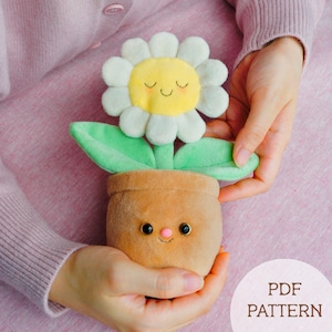 May include: A plush flower in a pot with a smiling face. The flower has white petals and a yellow center. The pot is brown, and the leaves are green. The text "PDF PATTERN" is at the bottom.