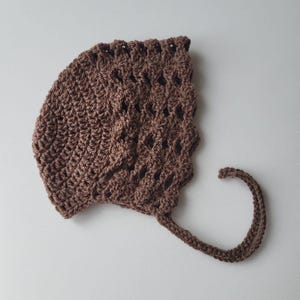 May include: A brown crocheted bonnet with a scalloped edge and a tie. The bonnet is made of a textured yarn and has an openwork design on the top. The bonnet is designed for a baby or toddler.