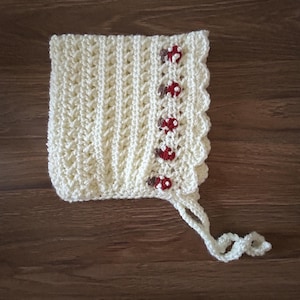 Puede incluir: Gorro de bebé de ganchillo color crema, con borde festoneado y detalles de setas rojas y blancas. El gorro tiene un patrón texturizado y se ata con una cuerda de ganchillo. Un accesorio hecho a mano y acogedor.