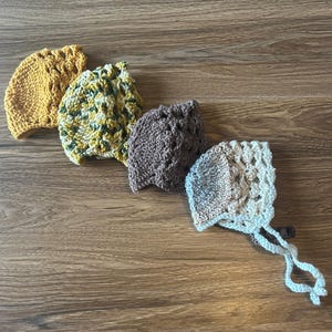May include: Four crocheted hats in different colours and patterns. The hats are made of yarn and have a variety of stitches. The hats are arranged in a row on a wooden surface.