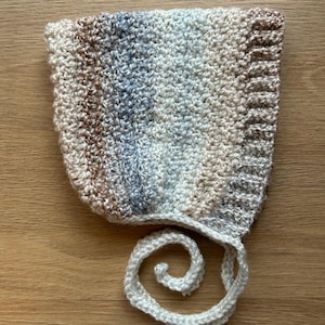 Peut inclure: Un bonnet de bébé fait à la main, crocheté, dans une palette de couleurs neutres crème, marron et bleu. Le bonnet présente un motif de point texturé et une attache délicate en spirale. Ce chapeau doux et confortable est parfait pour les nouveau-nés.