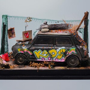 May include: A miniature black car with graffiti art, featuring yellow, pink, and green lettering. The model car is on a wooden base, with a backdrop of a green mesh fence and small paper cutouts. A small stack of rocks sits on the car's roof.