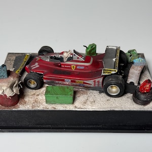 Puede incluir: Un modelo de coche de carreras rojo, con el número 11, sobre una base de diorama. El coche tiene neumáticos negros y un alerón plateado. El diorama incluye herramientas y accesorios en miniatura.