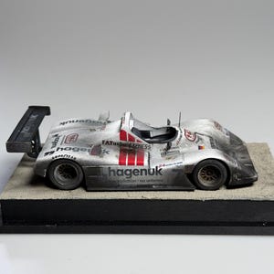 Könnte beinhalten: Silbernes Rennwagenmodell mit roten Streifen und Sponsorenlogos, darunter "hagenuk" und "FATurbo Express". Das Modell steht auf einem rechteckigen Sockel mit schwarzem Rand. Ein Heckflügel ist zu sehen.