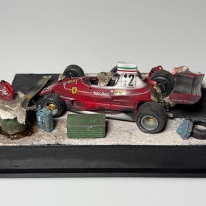 Op de afbeelding: Gedetailleerd model van een rode en witte vintage raceauto met nummer 12 op de helm. De auto staat op een diorama-basis met miniatuuraccessoires zoals brandstofblikken en een gereedschapskist.