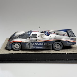 Könnte beinhalten: Ein detailliertes Modell eines Porsche-Rennwagens, hauptsächlich silberfarben mit blauen und roten Akzenten. Das Auto hat die Nummer 1, Rennaufkleber und das Wort "PORSCHE" auf der Seite. Es steht auf einem rechteckigen Sockel.