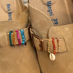 Könnte beinhalten: Ein Paar beigefarbene Wildleder-Slipper mit dem Markennamen Meik Mangni und der Größe 39 auf dem Fußbett. Die verstellbaren Riemen sind mit bunten Perlenringen verziert, darunter blaue Schutzaugen-Perlen und ein weißer Kaurimuschel-Anhänger.