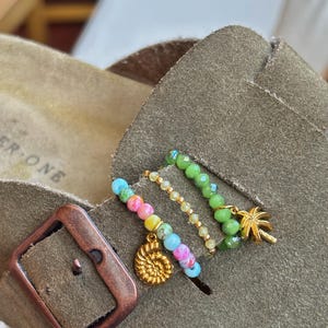 Op de afbeelding: Drie kralenringen bevestigd aan een suède sandalenband. Eén ring heeft veelkleurige pastelkleurige kralen met een gouden ammonietbedel. De tweede is een dunne band van kleine gele kralen. De derde heeft groene gefacetteerde kralen en een gouden palmboombedel.