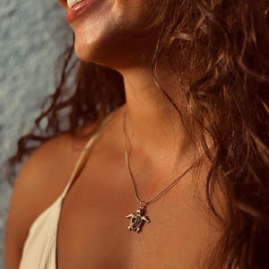 May include: A gold-colored turtle pendant necklace with a delicate chain. The turtle pendant has a dark center and is worn against a light-colored top. The image is a close-up, showing the necklace and part of the person's face.