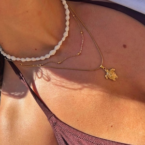Può includere: Un set di collane a strati con una collana di perline di conchiglie bianche, una delicata catena d'oro con piccole perle e un ciondolo a forma di tartaruga dorata. Le collane sono indossate sopra un top bikini bordeaux.