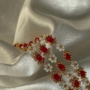 Peut inclure: Un bracelet en perles avec des perles rouges et dorées disposées en motif floral. Le bracelet est sur un fond de tissu blanc.